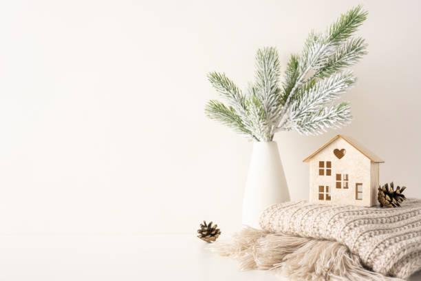 istock Minimalist side view Scandinavian style decor featuring a vase with snowy pine branches, a small wooden house, pine cones, and a cozy knitted blanket, evoking a warm and festive winter atmosphere 2188235965