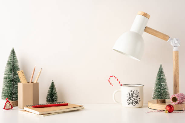 A side view minimalist winter workplace setup featuring Christmas-themed decorations, including a desk lamp, small trees, and holiday stationery. The scene evokes a cozy and festive Scandinavian style