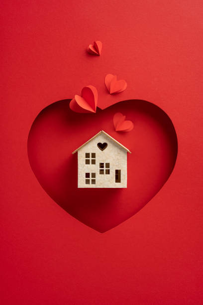 A vertical small house model with a heart window is displayed on a heart shaped red background, surrounded by paper hearts. Perfect for themes of love, home, and Valentine's Day