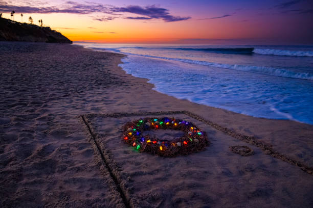 This is a creative photo of a Christmas wreath made out of seaweed with seashells and Christmas lights wrapped within it. There is a door drawn in the sand with a stick as the water is coming up close to it. This photograph was taken in Encinitas at Moonlight beach in San Diego county early in the morning capturing a beautiful sunrise.