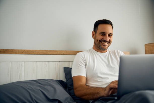 istock Happy man using laptop while relaxing in bed at home. 2188240070