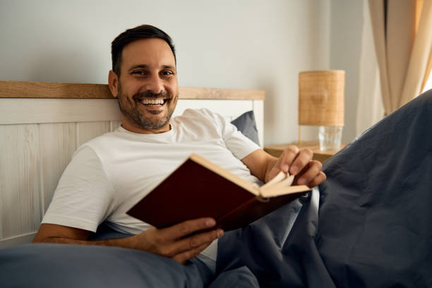 istock Happy man reading a novel while relaxing in bedroom and looking at camera. 2188240275