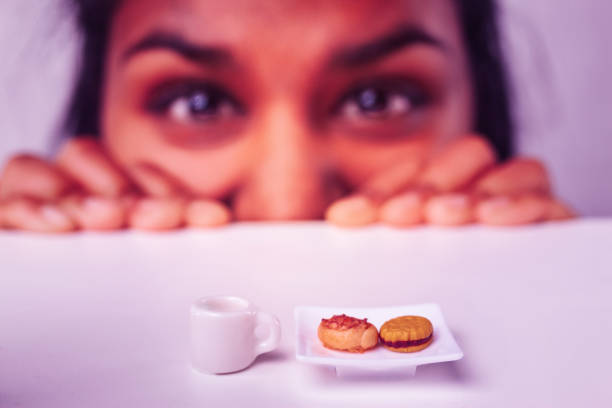 Young woman peeking over a table, eyes wide with curiosity, focused on a miniature plate of tiny cookies and a tiny cup of coffee, illustrating microscopic portions