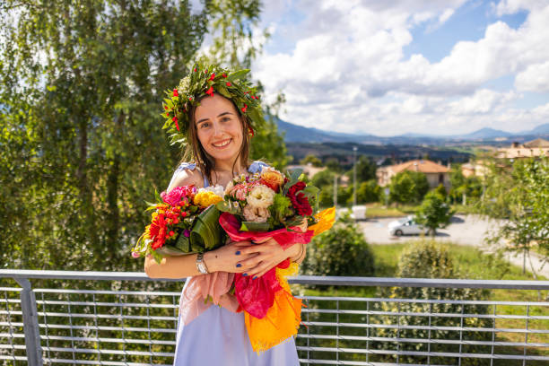Happy graduate in a light blue dress standing outside the ceremony venue, holding  a bouquets of flowers, wearing laurel wreath on her head