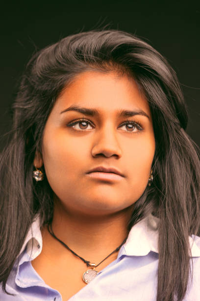 Portrait of a young indian woman with long dark hair and brown eyes, wearing a light blue shirt and a necklace, looking up with a serious and thoughtful expression against a dark background