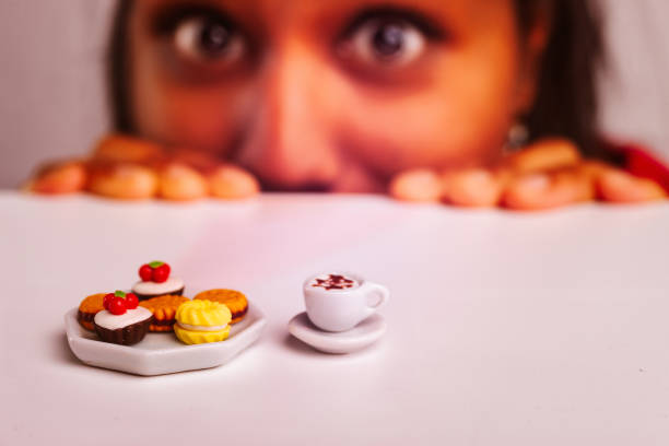 Woman is looking with curiosity at a miniature breakfast set with tiny cakes and a cup of coffee, highlighting the concept of microscopic portions and diet