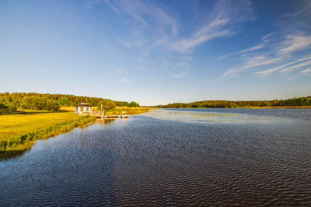 istock Peaceful river landscape with wooden dock, green grassy shoreline, and distant forest under clear blue sky. 2188264528