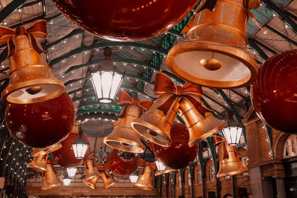 Christmas-themed decorations featuring oversized golden bells with red bows and bright red baubles hang from the ceiling at Covent Garden Market in London, UK. The festive scene showcases the iconic market's holiday transformation, with glittering ornaments and traditional lighting creating a warm and celebratory atmosphere. Captured during the 2024 holiday season, this image highlights the popular destination's charm and seasonal appeal.