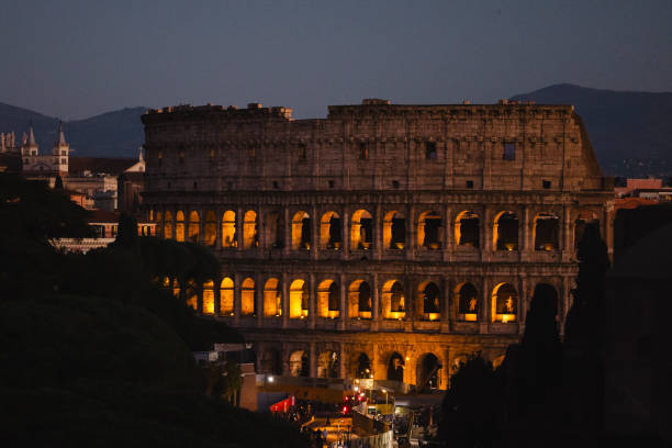 A wide angle colour photograph of a woman with her back to camera and with her pet scottie dog stands looking at the colosseum in Rome at night, ancient stone in the foreground,  trees and the arches of the historic place glowing as backdrop.