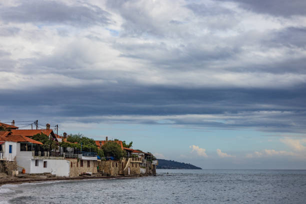 A serene coastal village with charming houses, a pebble beach, and dramatic clouds overhead. A paddle boarder adds a sense of adventure to the peaceful scene.