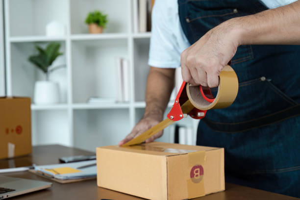 istock Man Sealing Cardboard Box with Tape in Home Office Setting for E-commerce or Small Business Packaging and Shipping 2188338816