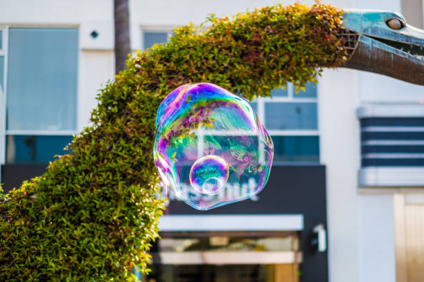 A single vibrant soap bubble showcases rainbow colors and light reflections, floating against an urban background with greenery. The dynamic play of light highlights its delicate, ephemeral beauty.