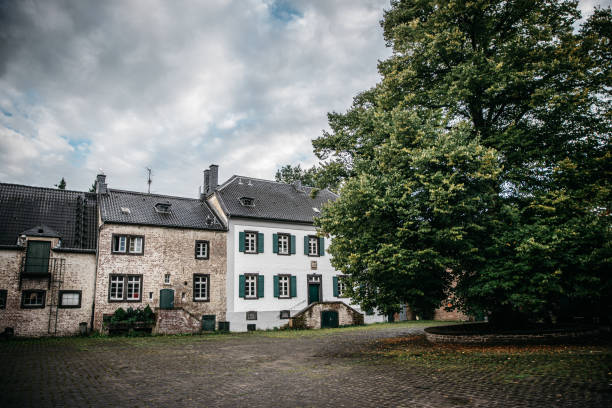 Historic residential buildings with characteristic German architecture featuring exposed brick and white-painted facades with green shutters. Traditional cobblestone courtyard surrounded by well-preserved townhouses under a dramatic cloudy sky, showcasing typical European architectural elements including dormer windows and steep tiled roofs.