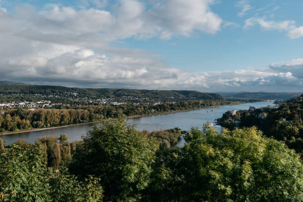 Scenic aerial view of the Rhine River Valley with rolling hills, small towns, and lush vegetation along the riverbanks. The image captures a dramatic sky with white clouds over the meandering river, surrounded by forested hillsides and residential settlements. A barge can be seen navigating the river, while historic architecture dots the landscape. The photograph was taken during daytime, showcasing the natural beauty and human settlement patterns typical of the Rhine region.