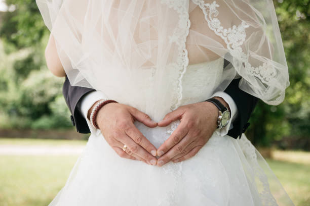 Close-up view of groom's hands forming a heart shape on bride's white lace wedding dress. The romantic gesture is captured in an outdoor garden setting with natural greenery in the background. The groom wears a formal black suit with white shirt and a brown leather bracelet, while sporting a wedding ring. The bride's dress features delicate lace details and tulle fabric, creating an elegant and timeless wedding photograph.