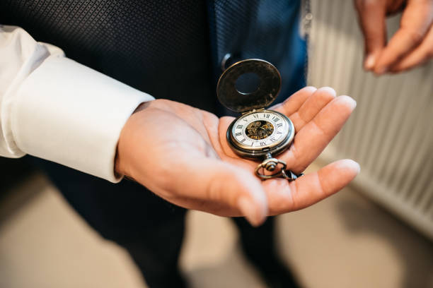 Close-up view of an elegant vintage pocket watch with exposed mechanical movement and roman numerals displayed in an open palm wearing a white dress shirt sleeve. The antique timepiece symbolizes precious moments and family heirlooms during a wedding celebration, creating a timeless and emotional detail shot perfect for wedding photography and storytelling.