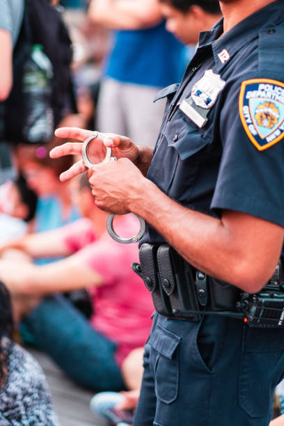 Police Officer Holding Handcuffs During a Law Enforcement Operation in a Public Setting, Demonstrating Authority and Readiness, Surrounded by a Seated Crowd Observing the Situation on a Busy Day.