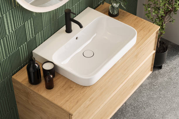 istock Close-up of a minimalist bathroom vanity with a white rectangular sink, wooden countertop, and green textured tile wall, styled with black soap dispensers and decorative plants. 2188360039