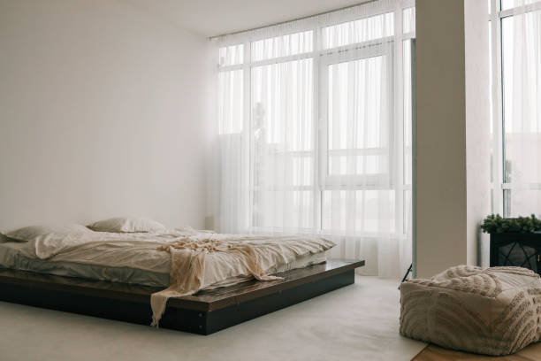 istock Minimalist bedroom with a cozy unmade bed, soft beige tones, and large windows draped with sheer curtains, letting in natural light. Tranquil and serene interior 2188370582