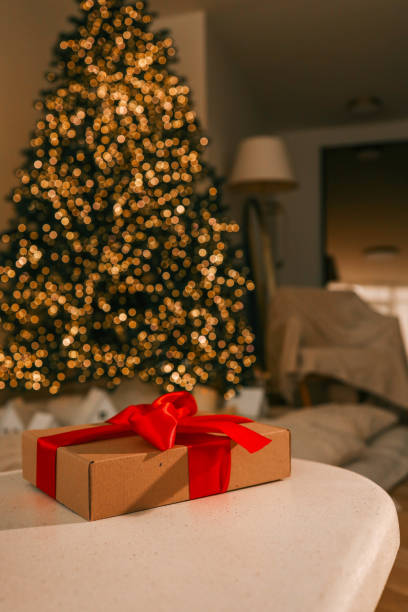 istock Festive Christmas scene with a wrapped gift box featuring a bright red ribbon on a table. A beautifully lit Christmas tree sparkles in the background, creating a warm holiday ambiance 2188370586