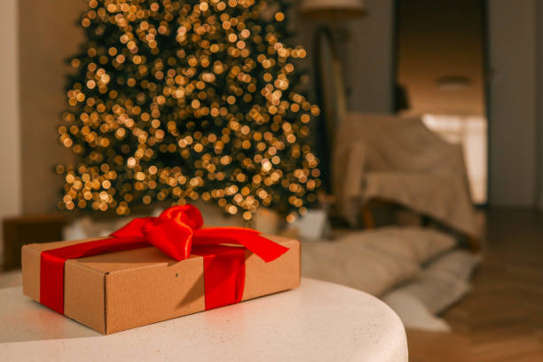 Festive Christmas scene with a wrapped gift box featuring a bright red ribbon on a table. A beautifully lit Christmas tree sparkles in the background, creating a warm holiday ambiance