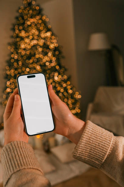 istock Person holding a smartphone with a blank screen in a cozy holiday setting. A decorated Christmas tree sparkles warmly in the background, creating a festive and serene atmosphere 2188370590