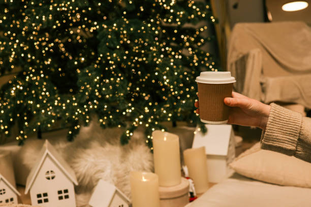 istock Hand holding a brown takeaway coffee cup with a white lid in a cozy holiday setting, featuring a lit Christmas tree, glowing candles, and decorative miniature houses in the background 2188371620
