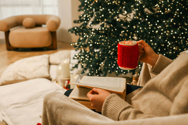 istock A cozy holiday scene featuring a person reading an open book while holding a festive red mug filled with marshmallows 2188371647