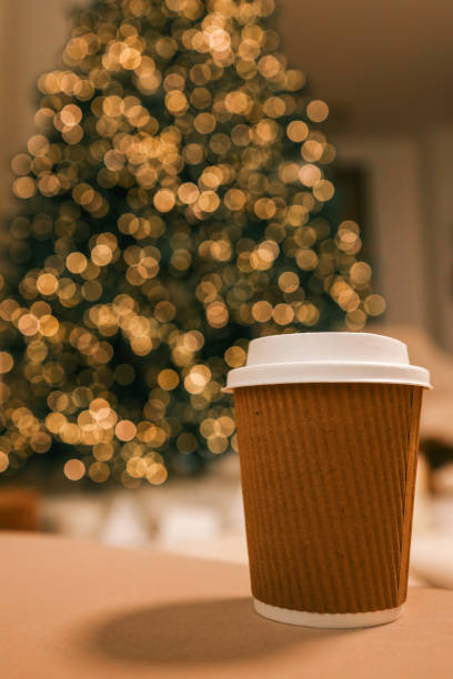 Takeaway coffee cup in eco-friendly brown paper holder with white lid, placed on a soft background with Christmas tree bokeh lights. Perfect for festive, cozy, and eco-conscious themes
