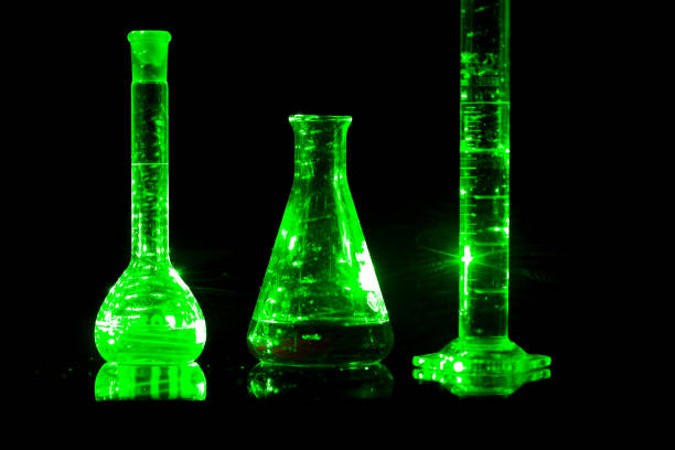Erlenmeyer flask, graduated cylinder and florence flask containing glowing green liquid, reflecting on a dark surface