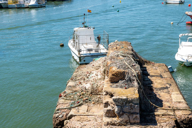 Isla Cristina, Huelva - Portugal - 09-09-2024: Old bridge pillar in water, covered with nets and ropes. In the background, fishing boats and storage buildings, with a blue sky above.