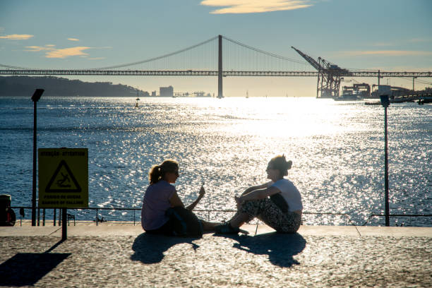 Lissabon, Lisboa - Portugal - 09-30-2024: Two women sit on a paved square with a view of the sparkling Tejo water, Ponte 25 de Abril bridge, harbor crane, and a warning sign.