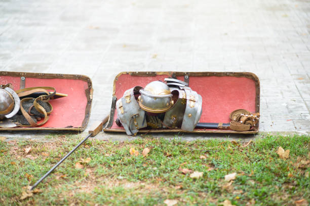 A display of Roman-style armor and red-painted shields laid out on the grass, including a detailed helmet with crest, metallic body armor, chainmail, and spear, representing ancient Roman military gear.