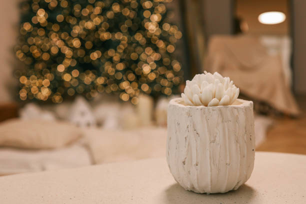 Elegant white decorative candle resembling a blooming flower in a textured white holder, placed on a table with festive bokeh lights and a blurred Christmas tree in the background