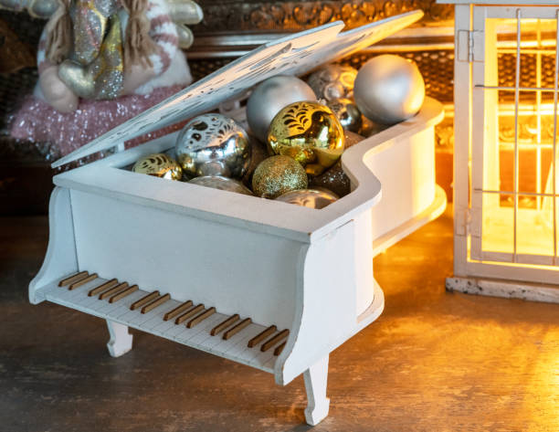 Festive Christmas decorations arranged inside a miniature white grand piano, featuring gold and silver ornaments. The warm glow of a lantern adds a cozy holiday atmosphere, seasonal and holiday-themed visuals