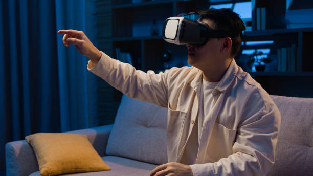 istock Asian adult man using modern technology explore virtual reality with 3d goggles eyeglasses playing game at night home Korean male wear VR glasses helmet play metaverse simulation at evening at couch 2188398020
