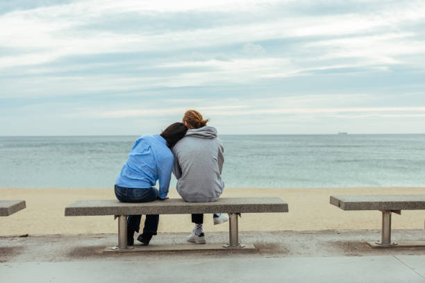 Two young adults find solace and connection as they sit together on a bench, gazing at the vastness of the sea