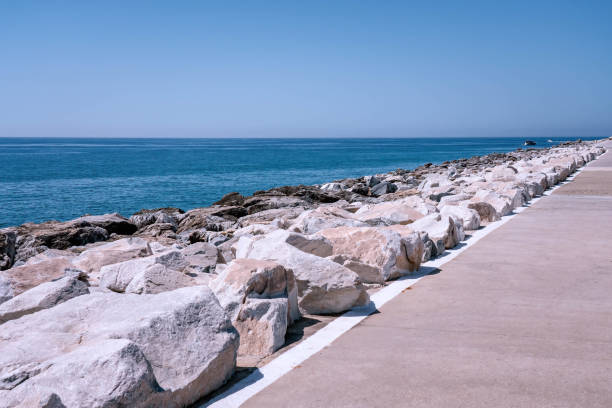 istock Large white rocks forming a breakwater, separating a paved walkway from the vast blue ocean 2188429490
