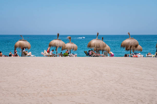 Tourists are enjoying a sunny day at the beach, relaxing under straw umbrellas and soaking up the sun's rays. PUERTO BANUS, MARBELLA - JULY 19 2022