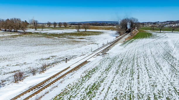 A vast expanse of snow-covered fields stretches across the rural landscape, with a railway track running alongside. The clear blue sky above enhances the winter scenery, revealing distant hills.