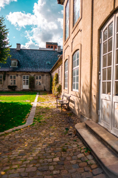 istock Charming Distinctive Architecture in a Sunlit Cobblestone Courtyard on a Bright Day with Blue Skies and White Clouds, Featuring an Inviting Wooden Bench and Green Grass, Creating a Tranquil Atmosphere 2188449306