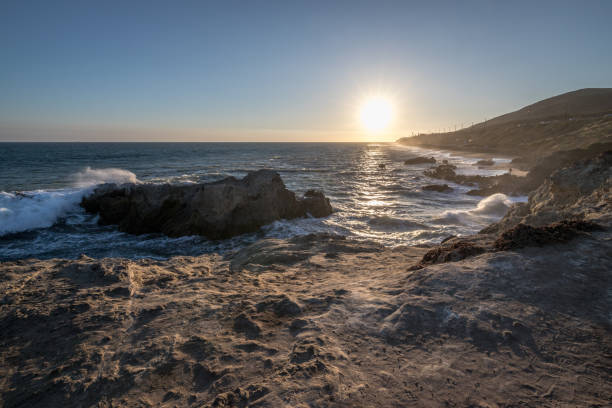 Waves crash against the rocks at Leo Carrillo State Beach as the sun sets, casting a warm glow over the coastal landscape in Malibu.
