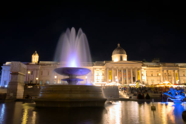 London, England - November 28, 2024: A winter's night in London, and here we see one of the ornate fountains found in Trafalgar Square. Beyond we see a brightly-lit Christmas Market and the National Gallery.