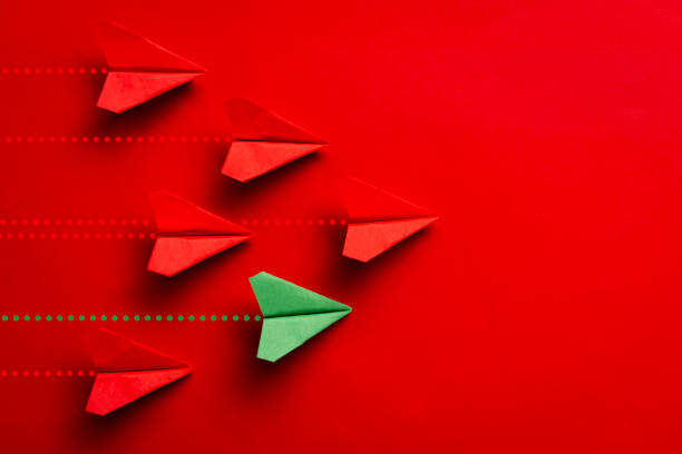 A green paper plane leading a group of red planes on a red background, symbolizing leadership, individuality, and standing out. The image highlights direction, strategy, and innovation.