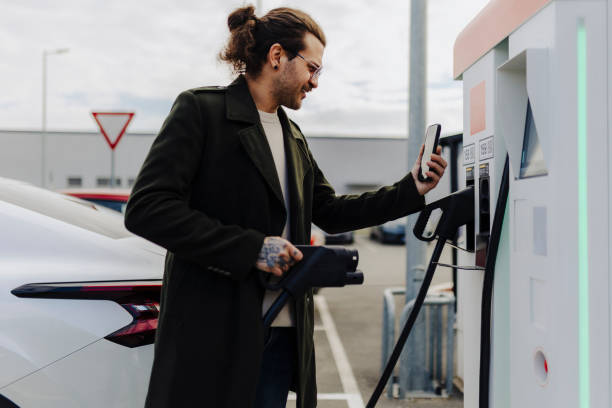 istock Man holding smartphone while charging car at electric vehicle charging station 2188533715