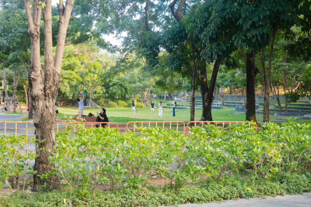 Relaxing thai people and students in Chulalongkorn University Centenary Park.. Captured from street Soi 26 at corner to main street Banthathong, in Bangkok  Pathum Wan. People are walking or sitting around, Some are using mobile phone. A group of female students is exercising with poles in artistic dance performance. At left side a woman is holding up a baby into air.
