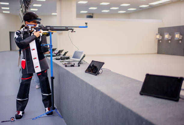 Precision Shooting Athlete at an Indoor Range A focused athlete practicing precision shooting in an indoor shooting range. The athlete is equipped with specialized gear, including a custom rifle, shooting jacket, and protective eyewear. The setting includes advanced digital target monitoring systems and training equipment, reflecting a professional training environment. Ideal for themes of sports, dedication, and high-performance training. military squad stock pictures, royalty-free photos & images