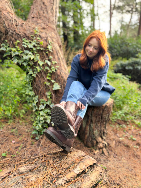 istock Image of attractive, redheaded woman wearing navy blue jumper, sitting on tree stump pulling leather walking boots on over jeans, woodland garden, ivy and pine needle ground cover, focus on foreground 2188560774