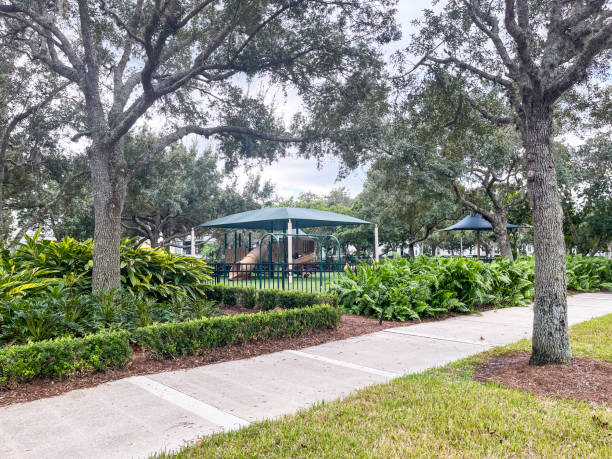 Sidewalk next to an urban playground located within an upscale neighborhood has a covered play area with real grass, surrounded by a nice fence,  and lined with oak trees and fine landscaping