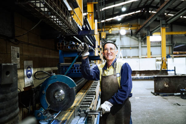 Worker in a factory setting adjusting a machine for processing metal materials in an industrial environment with safety gear. Blue collar worker cutting iron bars, looking at camera and smiling